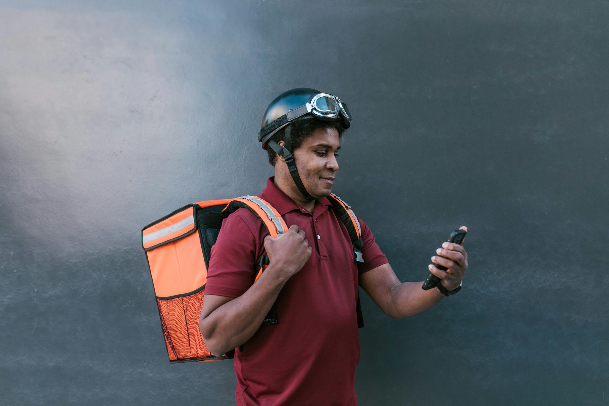 A delivery courier wearing a helmet and backpack checks his smartphone against a dark background.
